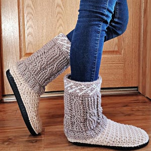 以下が含まれることがあります： ベージュとグレーの色合いの手編みスリッパブーツ。シャフト上部にケーブル編みのデザイン、カフ部分に白とグレーのパターンが施されています。ブーツは黒いソールで、室内での着用を想定してデザインされています。