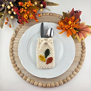 Op de afbeelding: Een herfsttafelsetting met een wit bord, bestek en een cr&egrave;mekleurige gebreide bestekhouder versierd met groene, gele en oranje bladeren. Het bord staat op een houten oplader met decoratieve kralen, aangevuld met herfstbladeren.