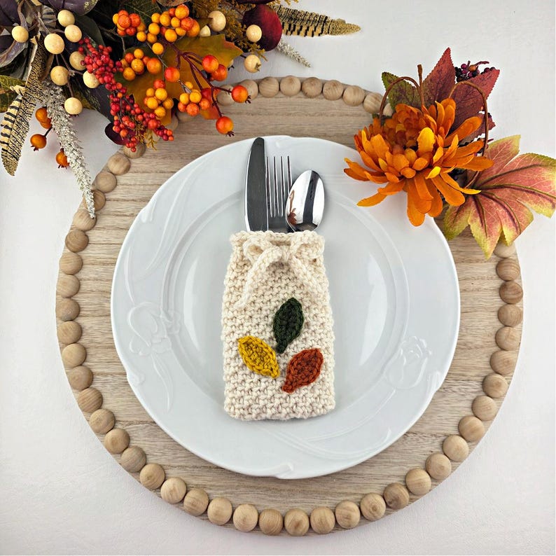 Op de afbeelding: Een herfsttafelsetting met een wit bord, bestek en een cr&egrave;mekleurige gebreide bestekhouder versierd met groene, gele en oranje bladeren. Het bord staat op een houten oplader met decoratieve kralen, aangevuld met herfstbladeren.