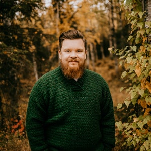 Pu&ograve; includere: Una persona indossa un maglione all'uncinetto verde scuro e pantaloni color cachi in una foresta. Il maglione ha un motivo strutturato e uno scollo a V. Lo sfondo presenta alberi con fogliame autunnale. Il testo "A Crocheted Simplicity" &egrave; in basso.
