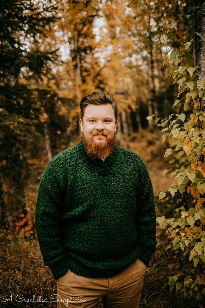 Pu&ograve; includere: Una persona indossa un maglione all'uncinetto verde scuro e pantaloni color cachi in una foresta. Il maglione ha un motivo strutturato e uno scollo a V. Lo sfondo presenta alberi con fogliame autunnale. Il testo "A Crocheted Simplicity" &egrave; in basso.