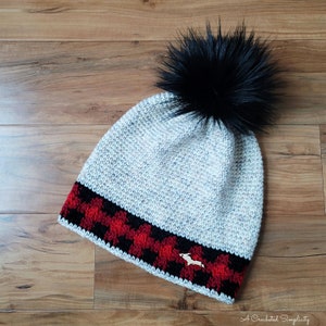 May include: A gray crocheted beanie with a black faux fur pom pom. The beanie has a red and black plaid trim with a small wooden moose charm.