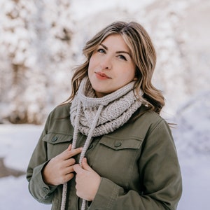 May include: A woman wearing a beige crocheted cowl neck scarf with long ties. The scarf is tied in a knot at the front of her neck. She is wearing a green jacket and is standing in a snowy landscape.