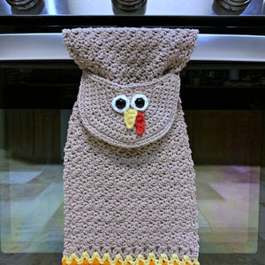 May include: A crocheted brown oven mitt shaped like a turkey with a red beak, yellow wattle, and two black eyes. The mitt has a colorful crocheted border in red, orange, yellow, and white.