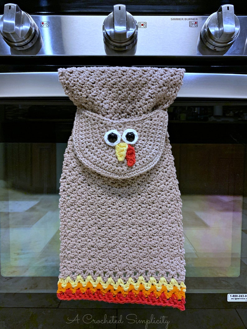 May include: A crocheted brown oven mitt shaped like a turkey with a red beak, yellow wattle, and two black eyes. The mitt has a colorful crocheted border in red, orange, yellow, and white.