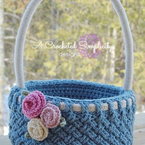 May include: A blue crocheted Easter basket with a white handle and a white and blue trim. The basket is decorated with three crocheted flowers in pink and white.