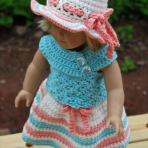 May include: A doll wearing a white, pink and blue crocheted hat, a blue crocheted top and a white, pink and blue striped crocheted skirt.