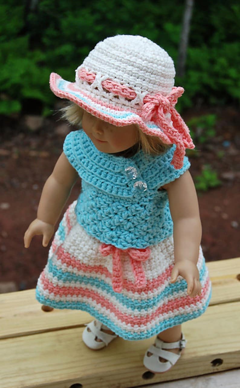 May include: A doll wearing a white, pink and blue crocheted hat, a blue crocheted top and a white, pink and blue striped crocheted skirt.