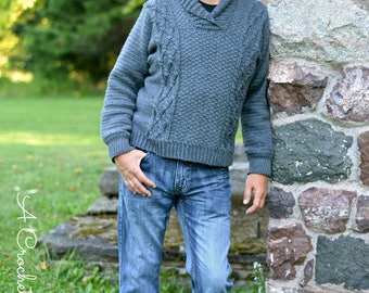 May include: A gray crocheted sweater with a cable knit pattern and a shawl collar. The sweater is worn by a person standing next to a stone wall.