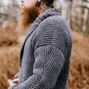 May include: A gray knitted cardigan with a textured stitch pattern. The cardigan has a collar and long sleeves. The cardigan is worn by a person with a beard and a tattoo on their neck.