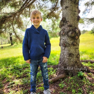 May include: A young boy wearing a blue crocheted sweater with a collar, distressed blue jeans, and gray sneakers. He is standing in a grassy area with a large tree behind him. The text "A Crocheted Simplicity" is visible in the lower right corner of the image.