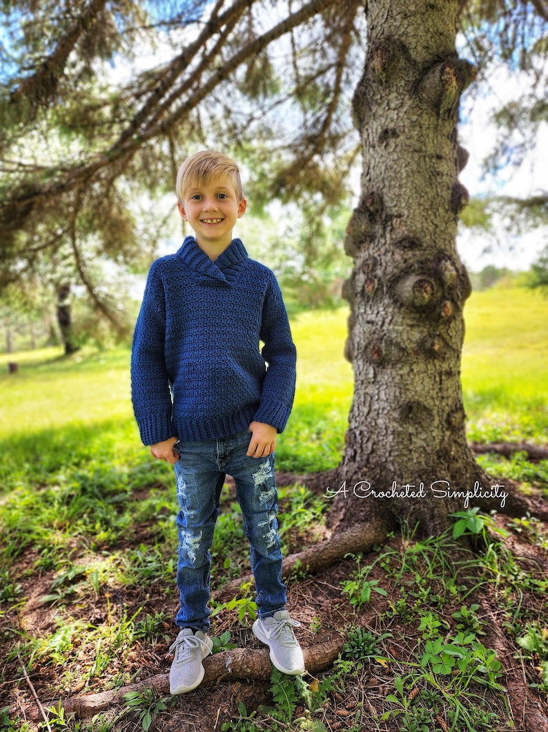 May include: A young boy wearing a blue crocheted sweater with a collar, distressed blue jeans, and gray sneakers. He is standing in a grassy area with a large tree behind him. The text "A Crocheted Simplicity" is visible in the lower right corner of the image.