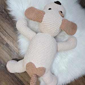 Puede incluir: Un peluche de perro hecho a mano con ganchillo en crema y marr&oacute;n claro. El perro tiene orejas marrones, una mancha marr&oacute;n en la cabeza y un parche en forma de coraz&oacute;n en la parte trasera. La cola es marr&oacute;n claro con una punta blanca. El peluche est&aacute; sobre una superficie blanca y esponjosa.