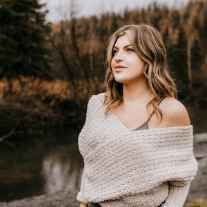 May include: A woman wearing a light beige crochet sweater with a draped neckline and long sleeves. She is standing in front of a body of water and a forest background.
