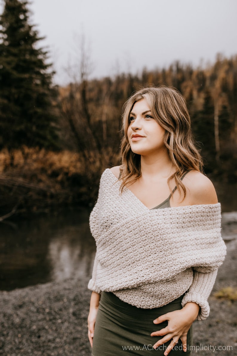 May include: A woman wearing a light beige crochet sweater with a draped neckline and long sleeves. She is standing in front of a body of water and a forest background.