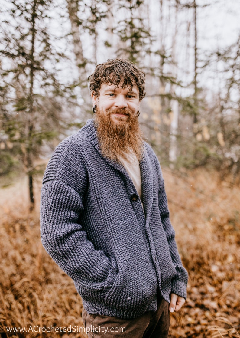 May include: A man wearing a blue knitted cardigan with a shawl collar and a single button closure. He is standing in a forest setting with brown leaves and trees in the background.