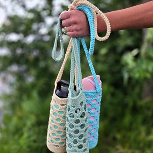 May include: Three crochet water bottle holders in beige, seafoam green, and light blue. Each holder has a matching strap and a decorative openwork design. The holders are designed to carry water bottles and are held in a hand against a blurred green background.