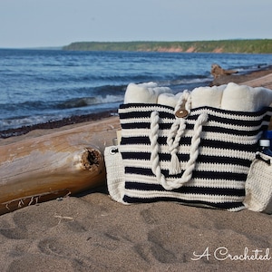 Può includere: Una borsa da spiaggia a righe in crochet con un manico in corda e una piccola borsa in crochet. La borsa è piena di asciugamani e una bottiglia d'acqua. La borsa è appoggiata su una spiaggia sabbiosa con un tronco in primo piano e un'acqua in lontananza.