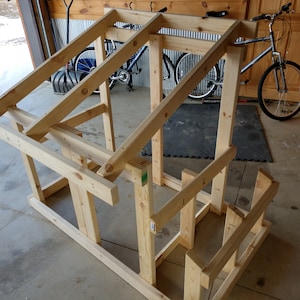 May include: A wooden frame for a small structure, possibly a birdhouse or a playhouse, is being assembled in a garage. The frame is made of unfinished pine wood and is supported by four legs. The frame is partially assembled, with the roof and walls still needing to be completed.