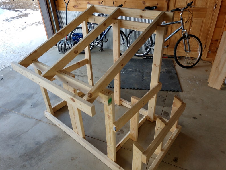 May include: A wooden frame for a small structure, possibly a birdhouse or a playhouse, is being assembled in a garage. The frame is made of unfinished pine wood and is supported by four legs. The frame is partially assembled, with the roof and walls still needing to be completed.