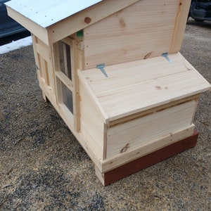 May include: A wooden chicken coop with a hinged roof and a small window. The coop is painted a light brown color and has a red base.