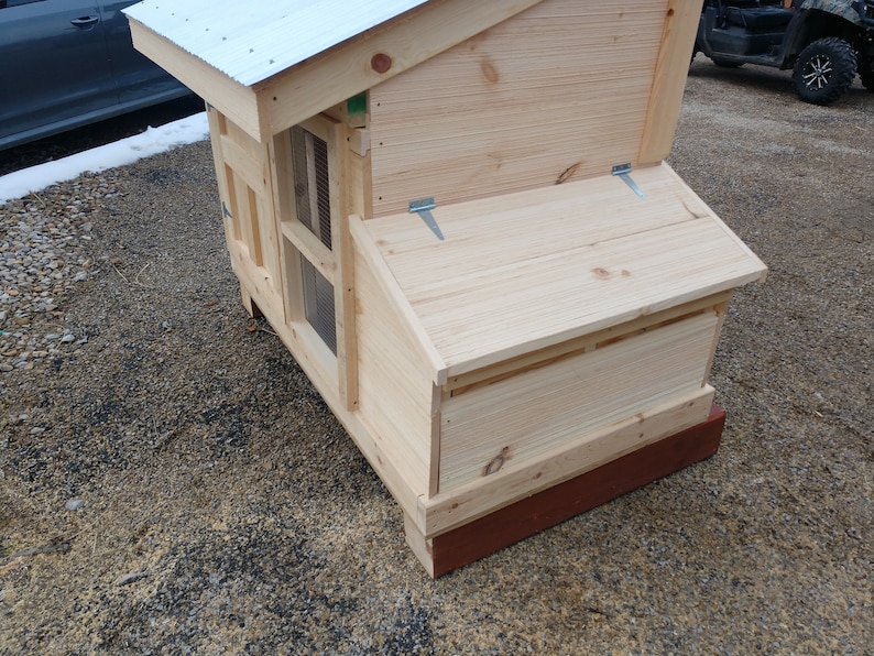 May include: A wooden chicken coop with a hinged roof and a small window. The coop is painted a light brown color and has a red base.