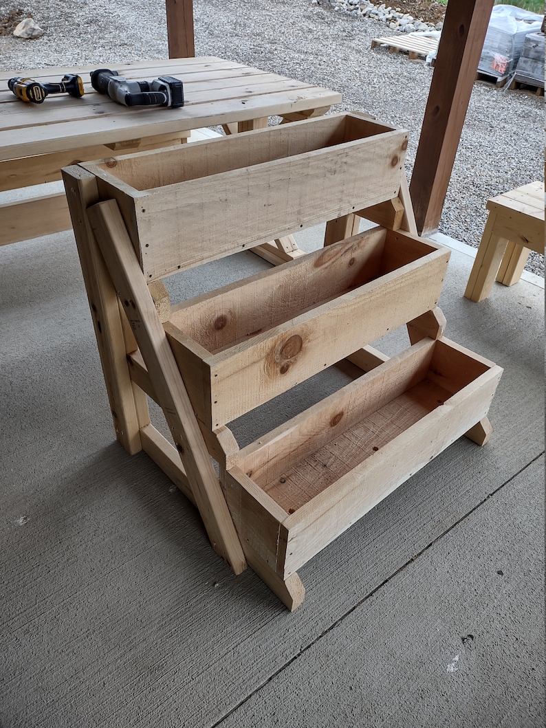 Puede incluir: Un soporte de plantas de madera con cuatro niveles, cada nivel es una caja rectangular. El soporte est&aacute; hecho de madera sin terminar y est&aacute; sentado sobre una superficie de concreto.
