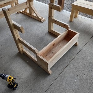 Puede incluir: Una jardinera de madera con respaldo y lados elevados, construida con tablones de madera sin terminar. La jardinera est&aacute; sentada sobre una superficie de concreto.