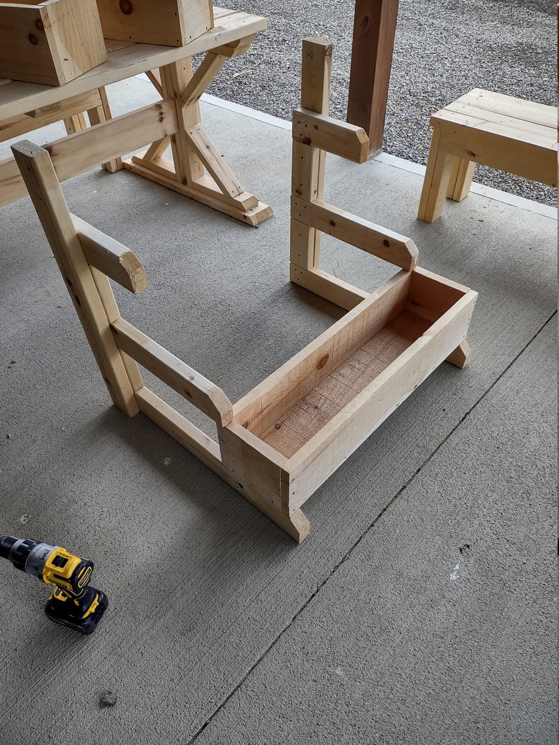 Puede incluir: Una jardinera de madera con respaldo y lados elevados, construida con tablones de madera sin terminar. La jardinera est&aacute; sentada sobre una superficie de concreto.