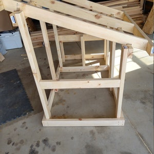 May include: A wooden frame structure with a sloped roof, constructed from unfinished pine lumber. The frame is standing on a concrete floor.