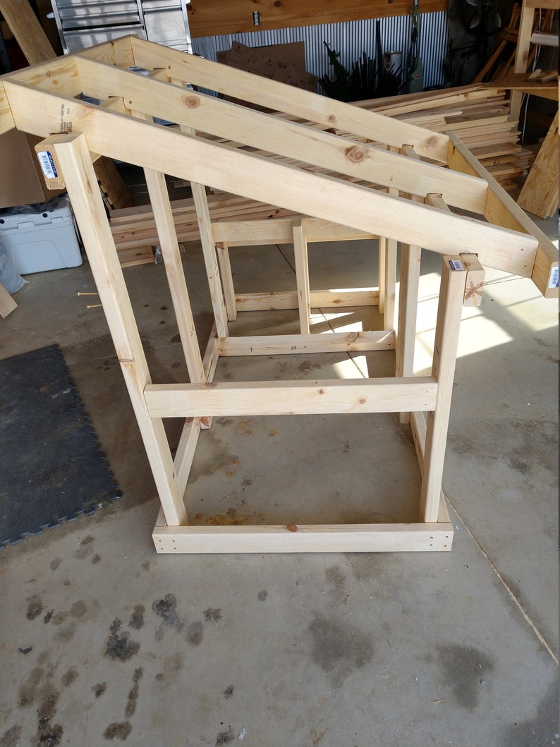 May include: A wooden frame structure with a sloped roof, constructed from unfinished pine lumber. The frame is standing on a concrete floor.