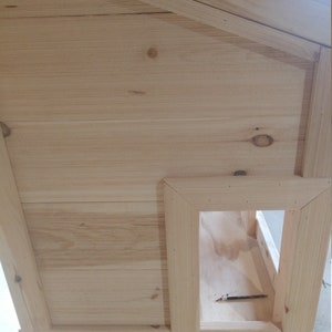 May include: A wooden dog house with a rectangular entrance. The house is made of light-colored wood and has a simple design.