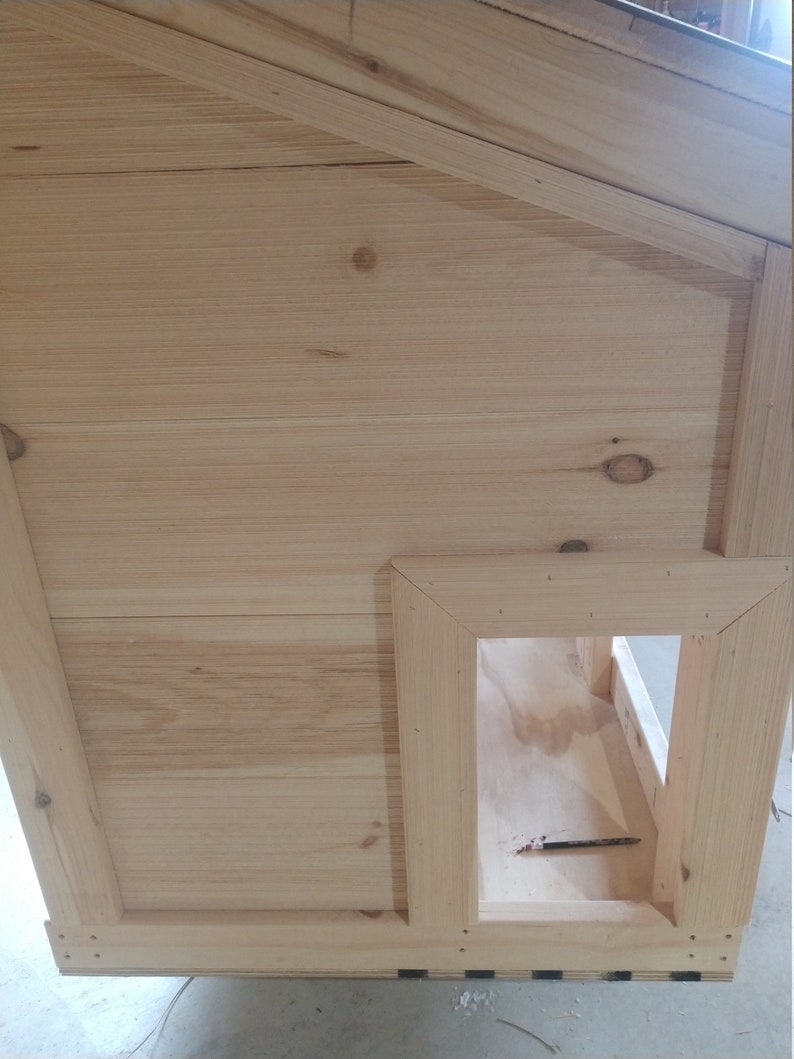 May include: A wooden dog house with a rectangular entrance. The house is made of light-colored wood and has a simple design.