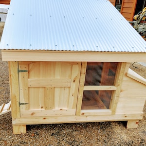 May include: A wooden chicken coop with a metal roof, a hinged door, and a wire mesh nesting box. The coop has a ramp leading up to the door.