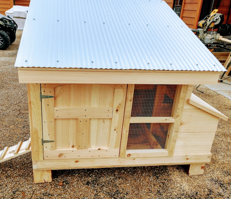 May include: A wooden chicken coop with a metal roof, a hinged door, and a wire mesh nesting box. The coop has a ramp leading up to the door.