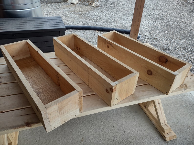Puede incluir: Cuatro jardineras de madera sin terminar, hechas de madera natural, est&aacute;n apiladas unas sobre otras en una superficie de madera.