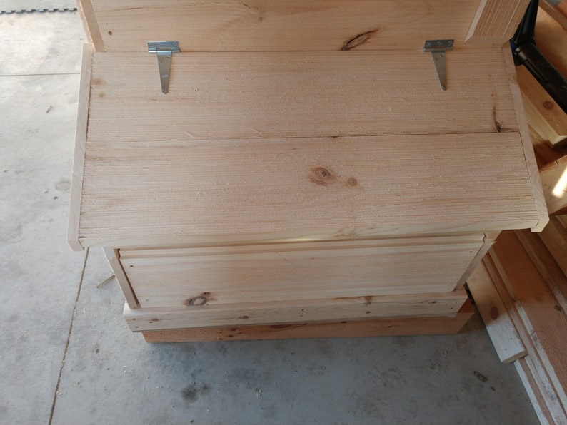 May include: A wooden storage box with a hinged lid and two metal hinges. The box is made of unfinished wood and has a simple design.