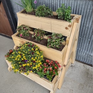 Puede incluir: Un macetero de madera en niveles con cuatro niveles, cada uno con diferentes plantas. El nivel superior tiene hierbas verdes, el segundo nivel tiene plantas de hoja roja y verde, el tercer nivel tiene flores amarillas y moradas, y el nivel inferior tiene flores rojas. El macetero est&aacute; hecho de madera natural y tiene un aspecto r&uacute;stico.