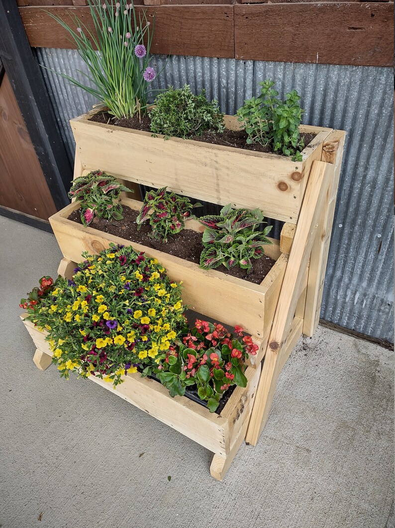 Puede incluir: Un macetero de madera en niveles con cuatro niveles, cada uno con diferentes plantas. El nivel superior tiene hierbas verdes, el segundo nivel tiene plantas de hoja roja y verde, el tercer nivel tiene flores amarillas y moradas, y el nivel inferior tiene flores rojas. El macetero est&aacute; hecho de madera natural y tiene un aspecto r&uacute;stico.