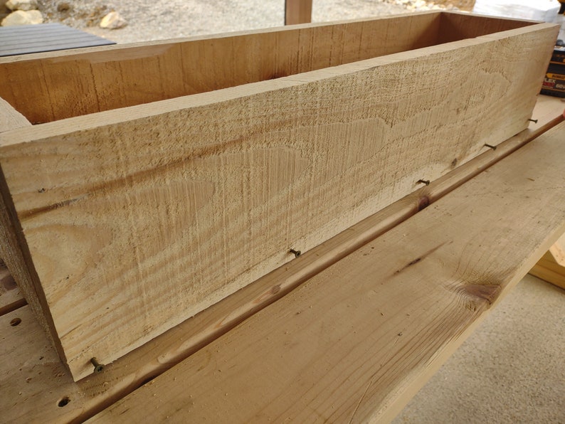 Puede incluir: Una caja de madera para plantar con un acabado natural. La caja se est&aacute; ensamblando con clavos. La caja est&aacute; sentada sobre una superficie de madera.