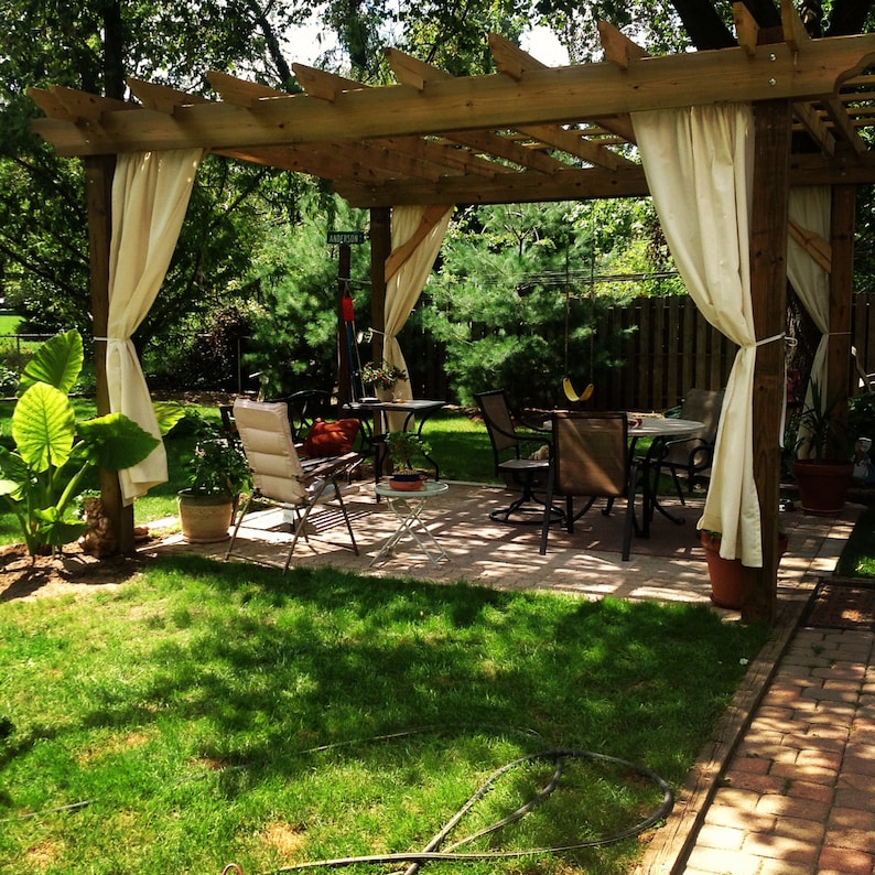 May include: A wooden pergola with white curtains, a patio set with chairs and a table, and green plants in pots. The pergola is in a backyard with a brick walkway.