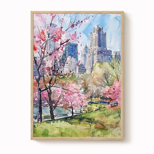 Op de afbeelding: Aquarel schilderij van een stadspark met bloeiende kersenbloesems. Het kunstwerk heeft een levendig kleurenpalet van roze, groen, blauw en bruin. Hoge gebouwen zijn zichtbaar op de achtergrond, omlijst door een lichtbruine houten lijst.