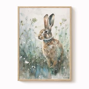 May include: A watercolour painting of a brown hare in a field of tall grass and wildflowers, framed in a light wood frame. The hare has long ears and a watchful gaze, set against a backdrop of soft, muted colours.