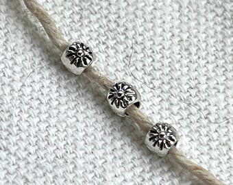 Puede incluir: Tres cuentas de metal plateado con un diseño de flor están enhebradas en un cordón blanco.