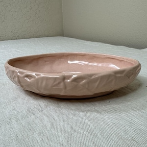 May include: A shallow, oval-shaped ceramic bowl in a soft, pale pink hue. The exterior surface features a textured, molded leaf pattern that wraps around the entire perimeter, while the interior has a smooth, glazed finish with visible crazing.
