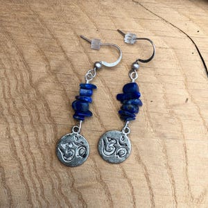 May include: A pair of silver-toned dangle earrings. Each earring features a strand of dark blue lapis lazuli chips and a circular charm with the Om symbol. The earrings have a hook closure.