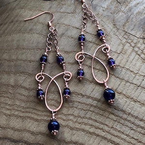 以下が含まれることがあります： 銅線で作られた、カスケードデザインのイヤリング。イヤリングは濃い紫色のビーズとチェーンのディテールが特徴です。イヤリングはフック留めです。イヤリングは木製の表面に展示されています。