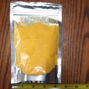 May include: A silver resealable pouch filled with a bright yellow powder. The pouch is sitting on a wooden surface.