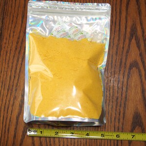 May include: A clear plastic bag with a silver iridescent finish contains a yellow powder. The bag is sealed with a zipper.