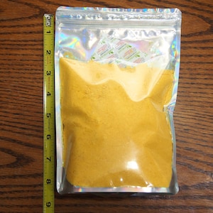 May include: A clear plastic bag with a silver iridescent finish contains a yellow powder. The bag is sealed with a zipper.